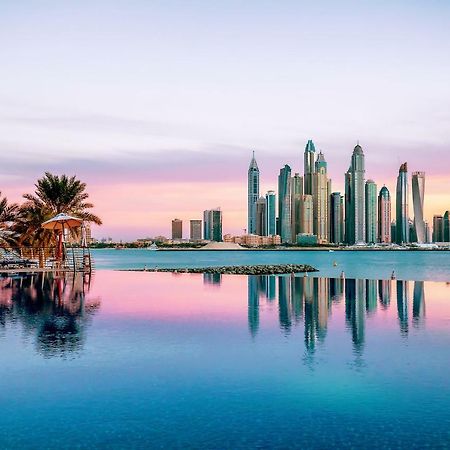 Dukes The Palm, A Royal Hideaway Hotel Dubai Exterior foto The image showcases a stunning skyline view of a modern city, likely during sunrise or sunset, as indicated by the beautiful pastel colors in the sky. The foreground features a calm body of water, reflecting the skyscrapers and the colorful sky. Lush