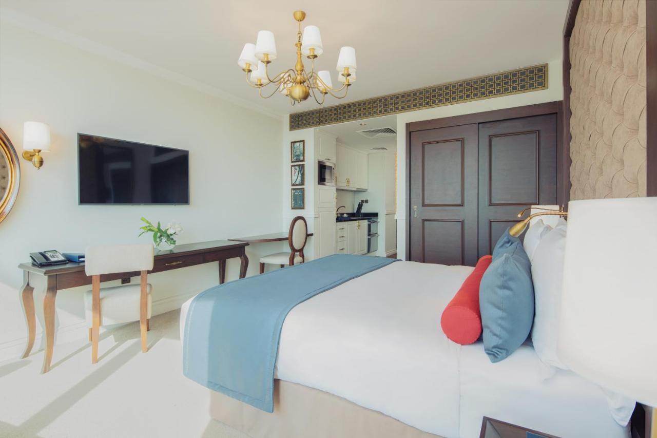 Dukes The Palm, A Royal Hideaway Hotel Dubai Exterior foto The photo depicts a modern hotel room featuring a neatly made queen-sized bed with decorative pillows in shades of blue and red. A stylish chandelier hangs from the ceiling, providing warm lighting. Opposite the bed, there is a wall-mounted flat-scre