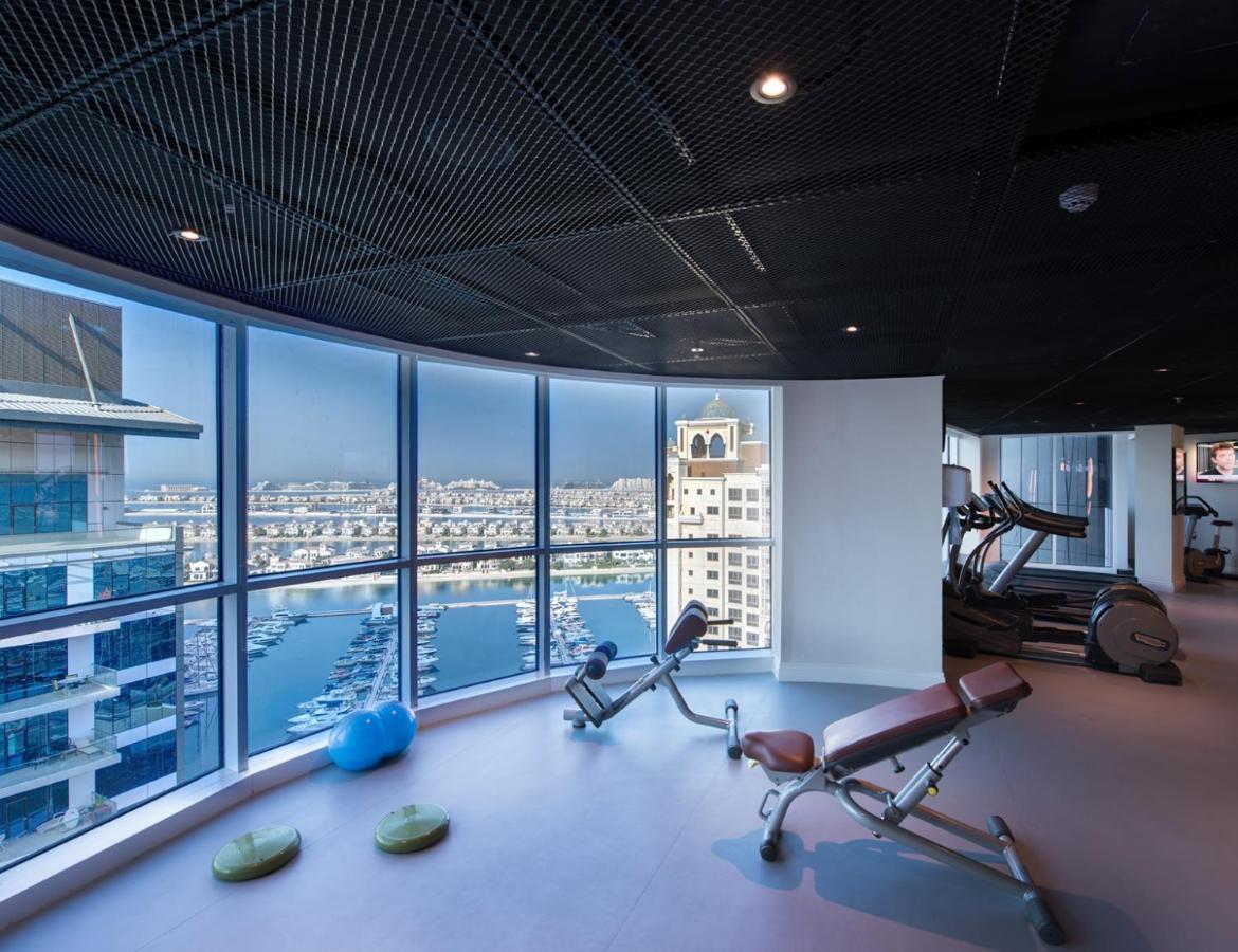 Dukes The Palm, A Royal Hideaway Hotel Dubai Exterior foto The photo shows a modern gym with large windows offering a panoramic view of a marina or waterfront area. Inside, there are exercise machines, including a couple of benches for weight training. The flooring is smooth, and there are a few exercise acc