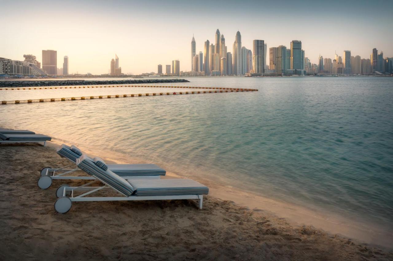 Dukes The Palm, A Royal Hideaway Hotel Dubai Exterior foto The photo shows a tranquil beach scene with several lounge chairs lined up on the sand, facing a calm body of water. In the background, a modern skyline is visible, featuring tall buildings and skyscrapers. The atmosphere appears serene, possibly dur