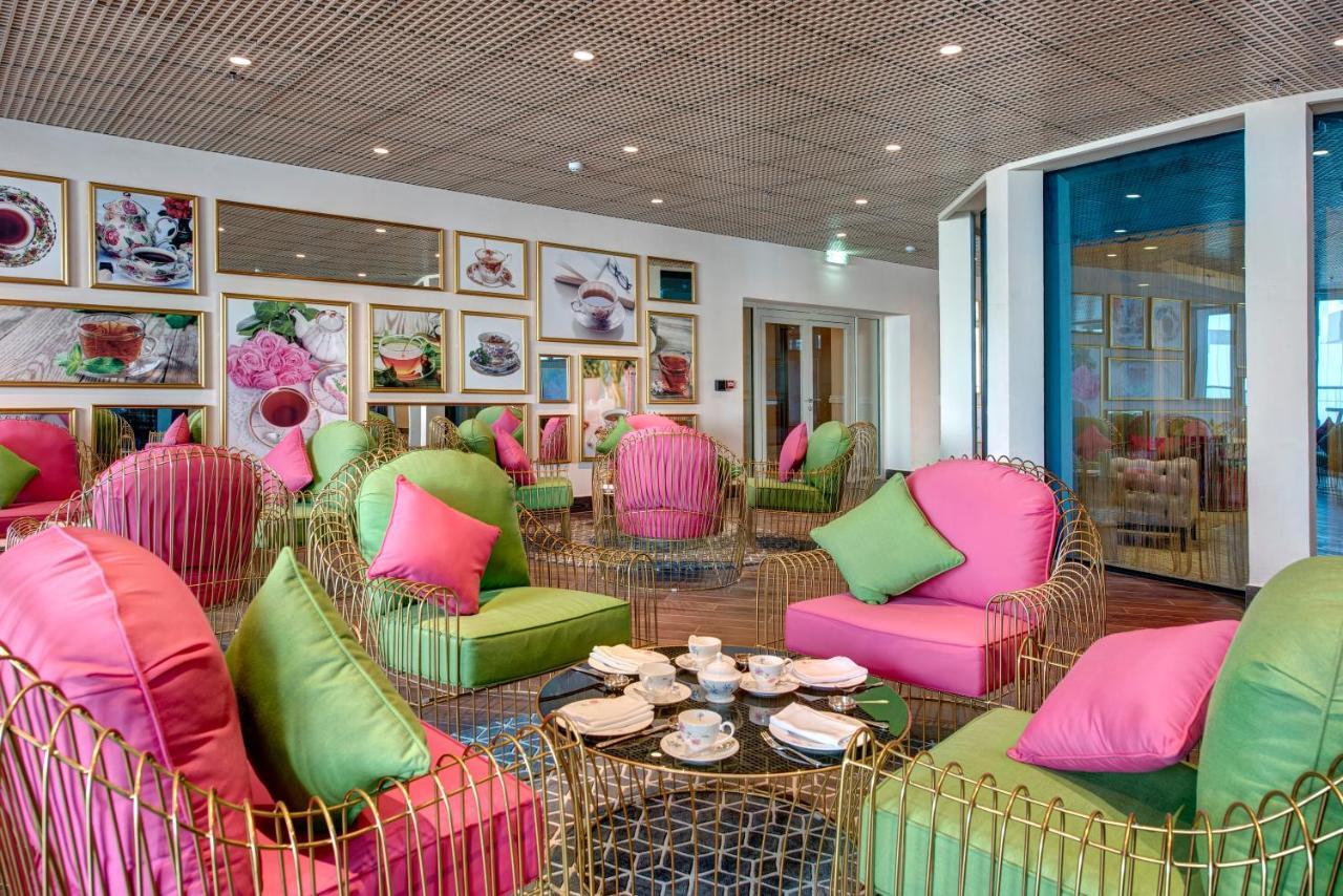 Dukes The Palm, A Royal Hideaway Hotel Dubai Exterior foto The photo shows a stylish and modern interior space, possibly a cafe or lounge. The seating area features elegant chairs with gold frames and vibrant cushions in shades of pink and green. There are several round tables with tea sets and small plates 