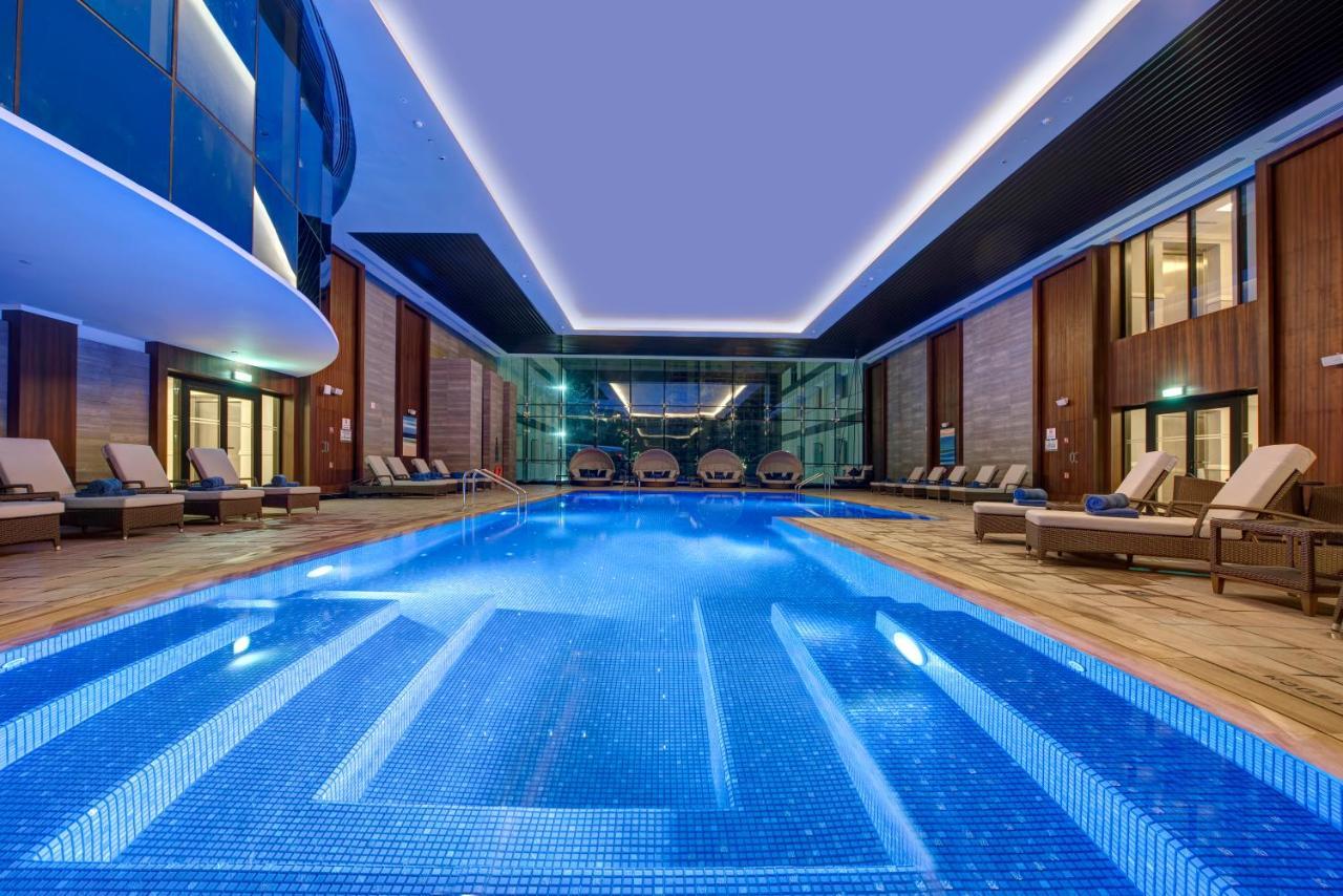 Dukes The Palm, A Royal Hideaway Hotel Dubai Exterior foto The photo shows a luxurious indoor swimming pool area. The pool is rectangular and features a blue tiled bottom, creating a vibrant hue in the water. Surrounding the pool are comfortable lounge chairs lined up along the edges. The area has large glas