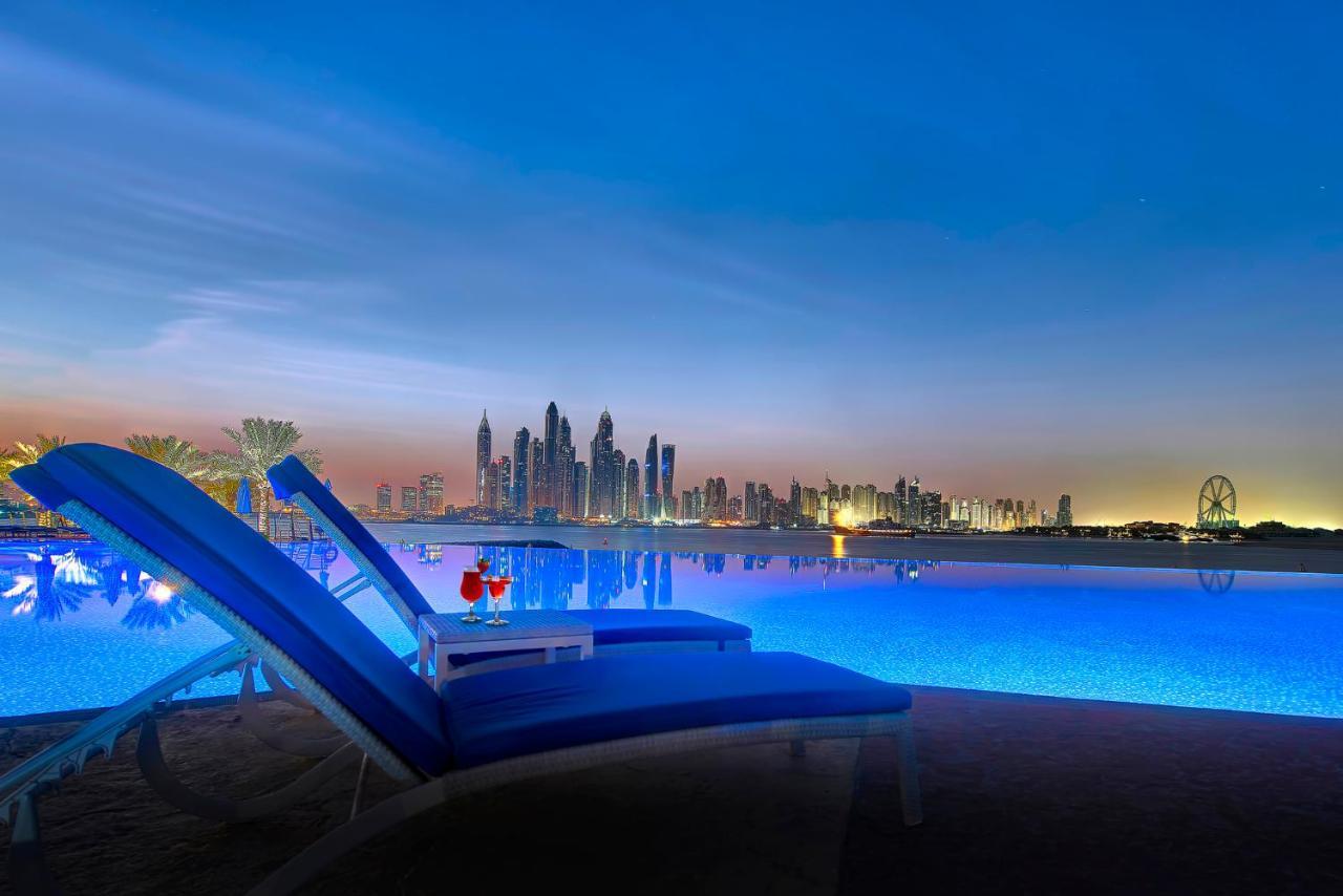 Dukes The Palm, A Royal Hideaway Hotel Dubai Exterior foto The image depicts a tranquil scene by a swimming pool, illuminated by soft blue lighting. In the foreground, there are two lounge chairs positioned beside the pool, suggesting a relaxing environment. A small table between the chairs holds two cocktai