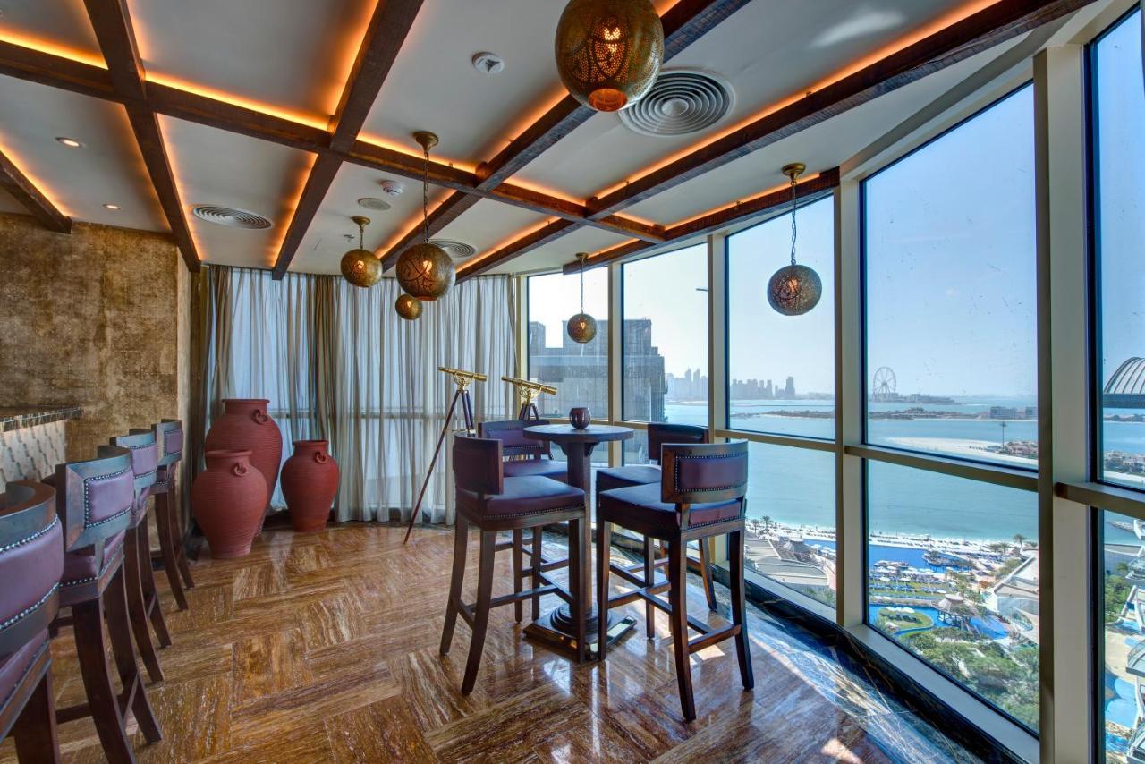 Dukes The Palm, A Royal Hideaway Hotel Dubai Exterior foto The photo depicts a modern, stylish interior of a restaurant or lounge. It features large windows that offer a view of a waterfront and cityscape. The design includes warm wooden beams and ambient lighting, creating a cozy atmosphere. There are sever