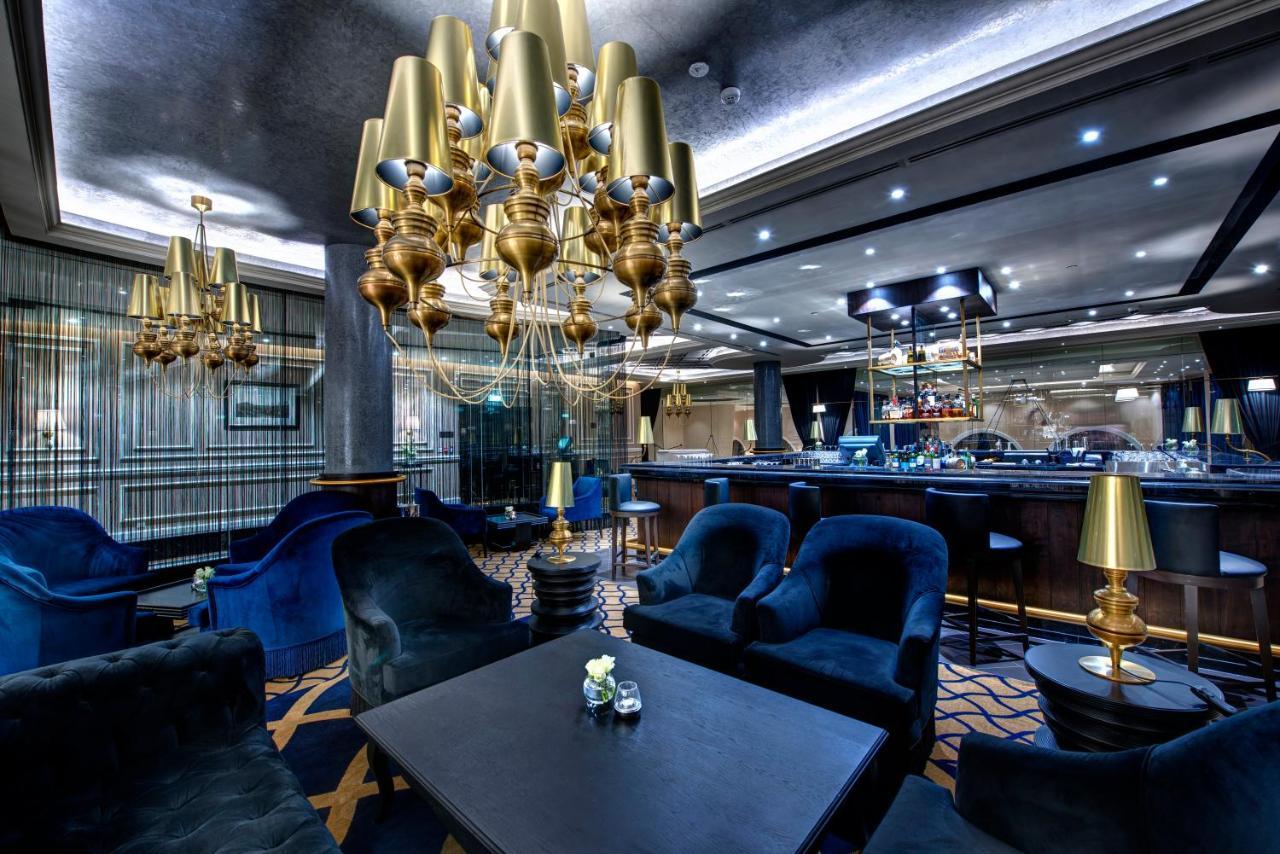 Dukes The Palm, A Royal Hideaway Hotel Dubai Exterior foto The photo displays an elegant interior of a lounge or bar area. It features plush blue seating, including armchairs around a dark table. A prominent chandelier with golden lamps hangs from the ceiling, providing ambient light. In the background, ther