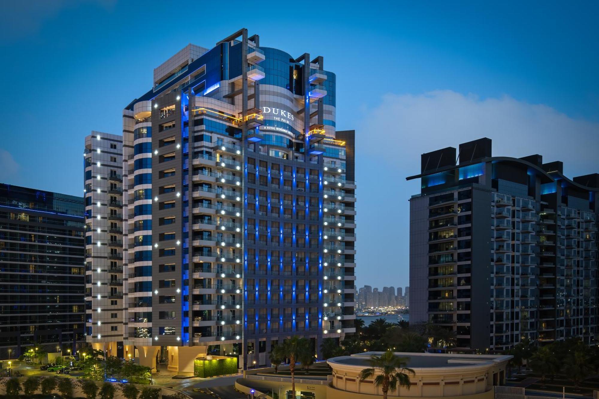 Dukes The Palm, A Royal Hideaway Hotel Dubai Exterior foto The photo shows a modern high-rise building, which appears to be a hotel or residential complex. The design features sleek lines and a glass façade, illuminated with blue lighting that enhances its contemporary look. Surrounding the main building are