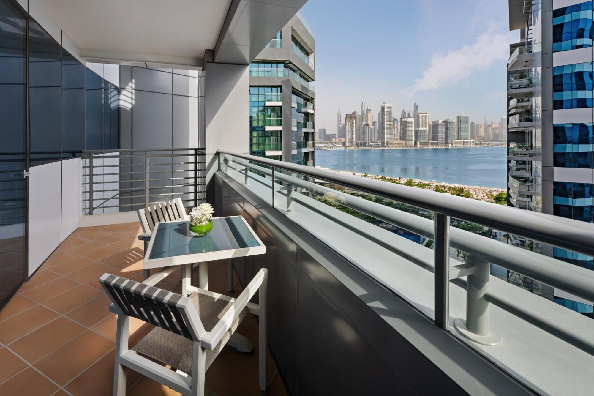 Dukes The Palm, A Royal Hideaway Hotel Dubai Exterior foto The photo shows a balcony with a modern design, featuring a small table and two white chairs. On the table, there is a vase with flowers. The balcony offers a view of a waterfront cityscape, with tall buildings in the background and a calm body of wa