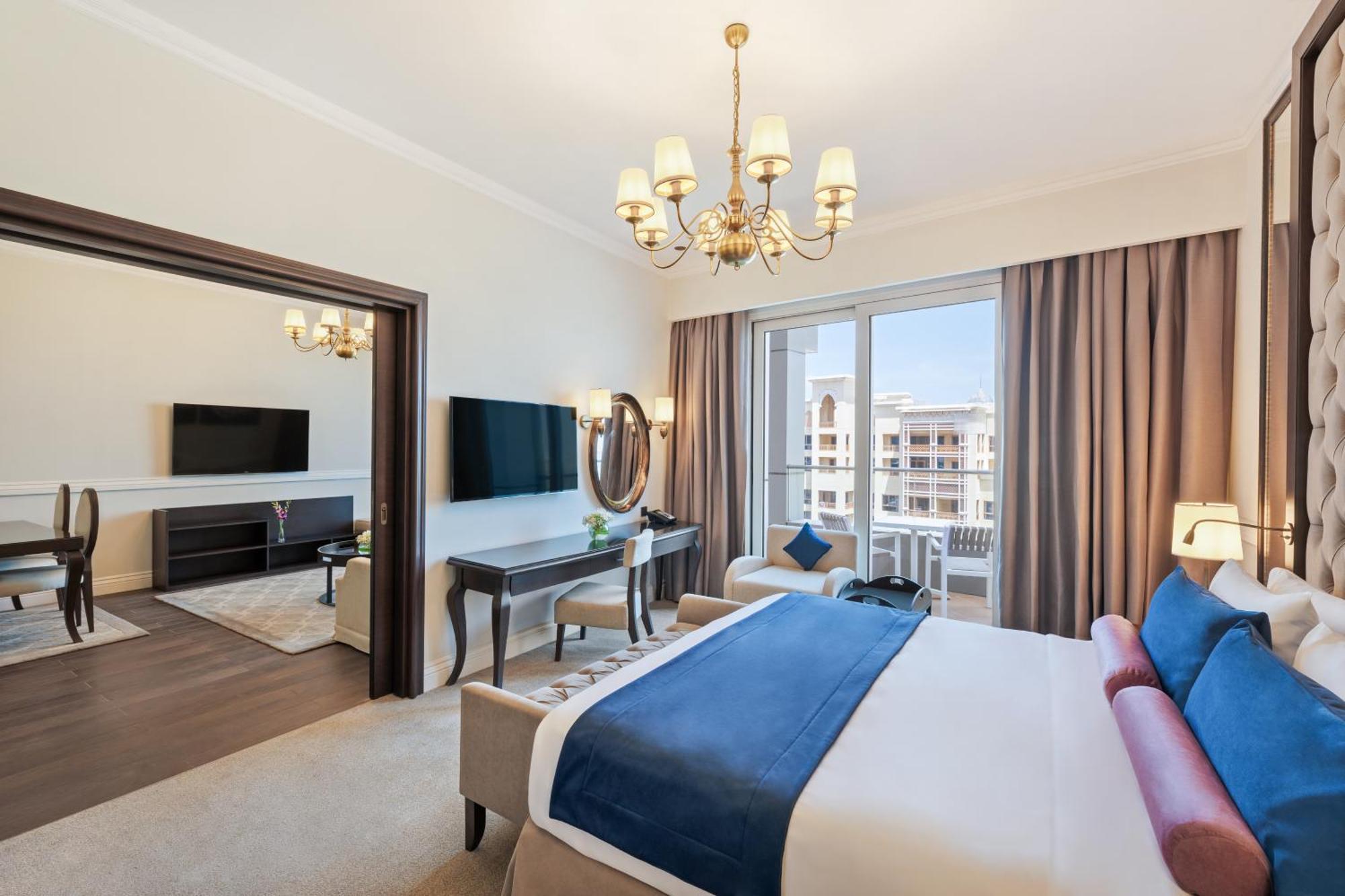 Dukes The Palm, A Royal Hideaway Hotel Dubai Exterior foto The photo depicts a stylish hotel room. It features a comfortable bed with blue and white bedding. The room includes a large window with curtains that allow natural light in, and there are two flat-screen TVs mounted on the walls. A chandelier hangs 
