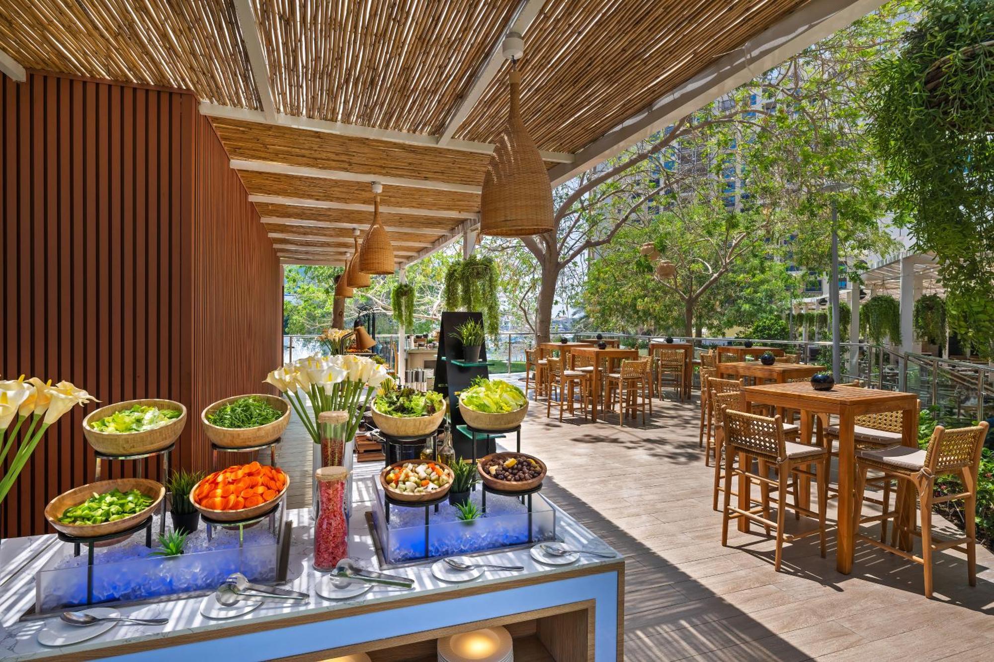 Dukes The Palm, A Royal Hideaway Hotel Dubai Exterior foto The photo showcases a bright and inviting outdoor dining space. In the foreground, there is a buffet table with various salads and fresh vegetables displayed in bowls, including items like lettuce, carrots, and other colorful ingredients, set against