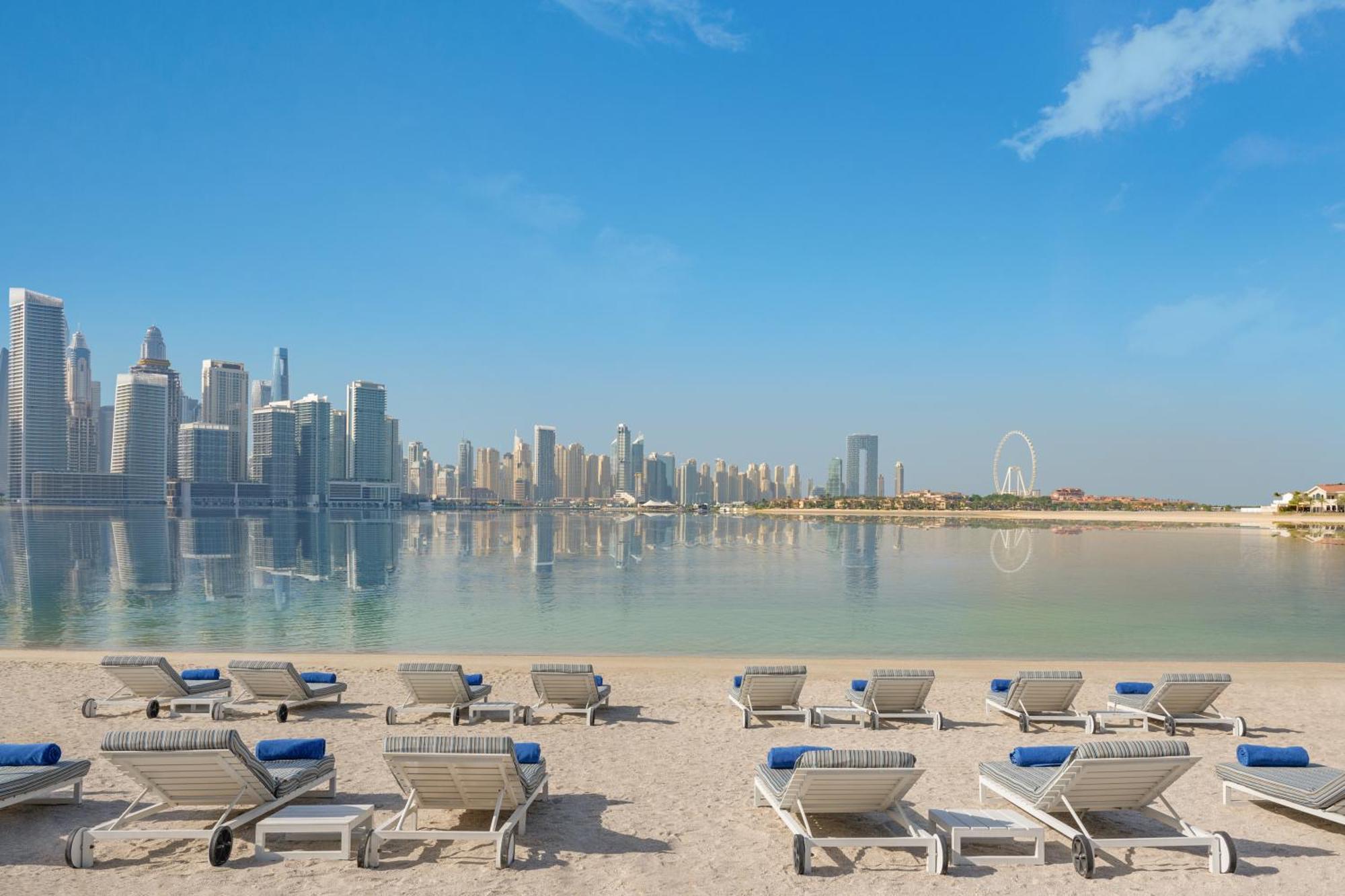 Dukes The Palm, A Royal Hideaway Hotel Dubai Exterior foto The photo shows a serene beach scene with several lounge chairs arranged in the foreground. These chairs are positioned on soft sand and have blue cushions. In the background, there is a stunning view of a modern skyline featuring tall buildings and 