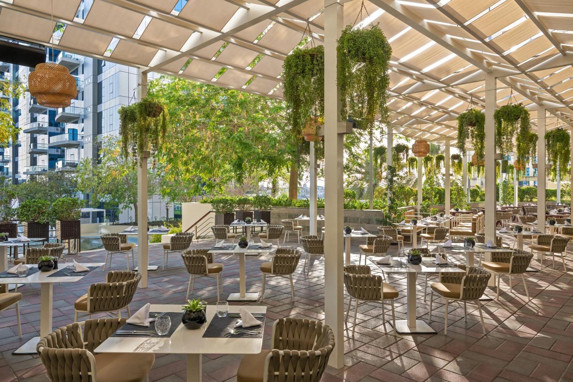 Dukes The Palm, A Royal Hideaway Hotel Dubai Exterior foto The photo shows a charming outdoor restaurant or café with a spacious seating area. The space is adorned with several plants hanging from the ceiling, creating a vibrant and lush atmosphere. There are rows of tables set with utensils and small decora