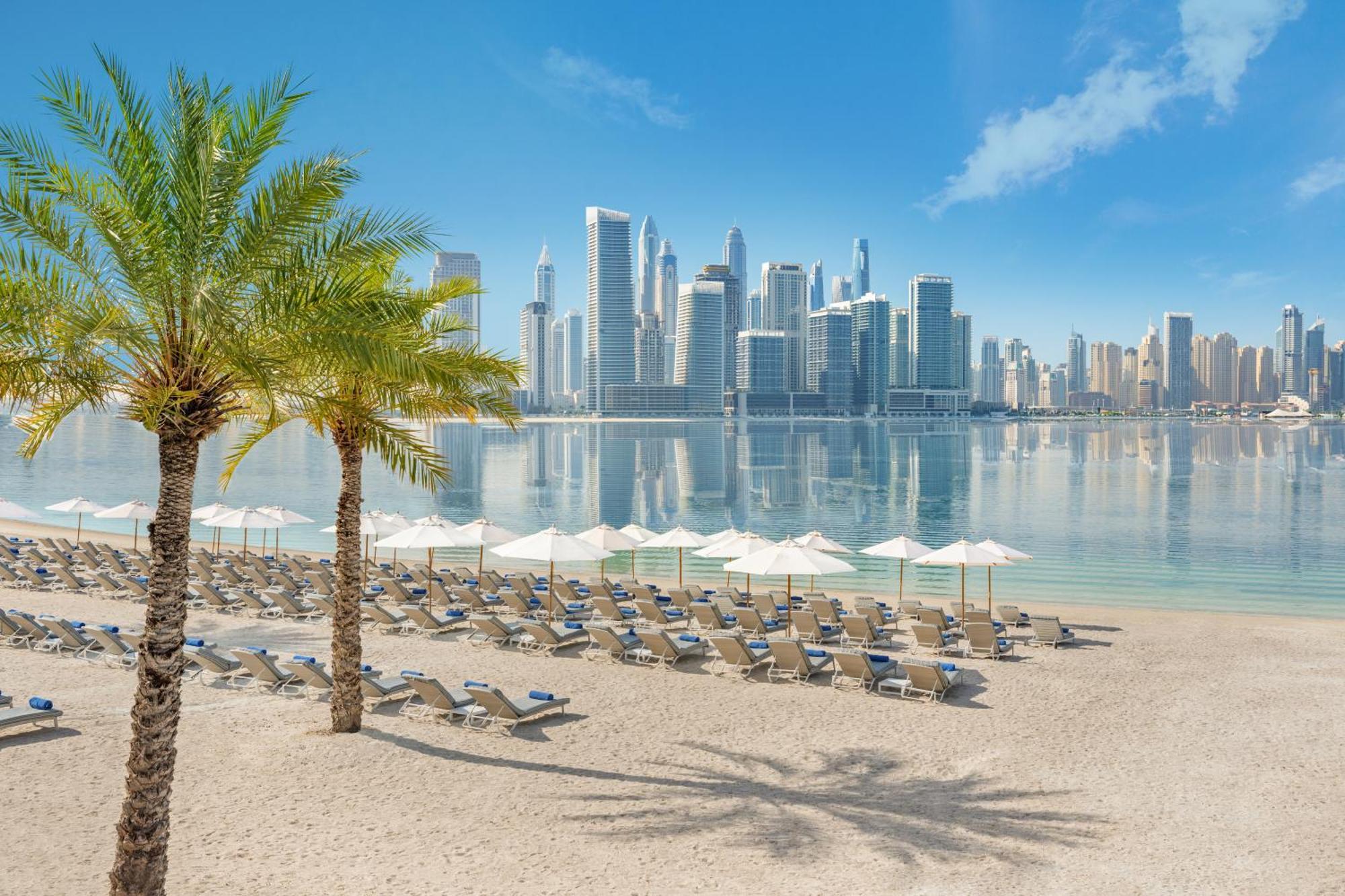 Dukes The Palm, A Royal Hideaway Hotel Dubai Exterior foto The photo depicts a tranquil beach scene with a view of a modern city skyline in the background. In the foreground, there are several lounge chairs arranged neatly on the sandy beach, accompanied by white umbrellas for shade. Palm trees add a tropica