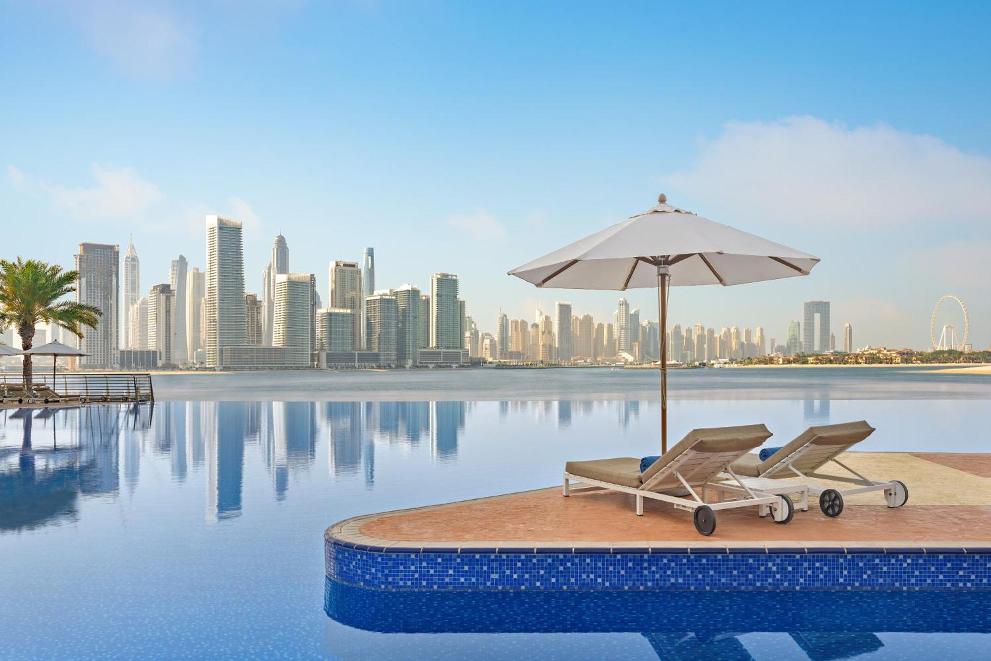Dukes The Palm, A Royal Hideaway Hotel Dubai Exterior foto The photo shows a serene and luxurious setting, featuring an infinity pool that seems to blend into a body of water. In the foreground, there are two lounge chairs positioned on a platform, accompanied by a large white umbrella providing shade. The b