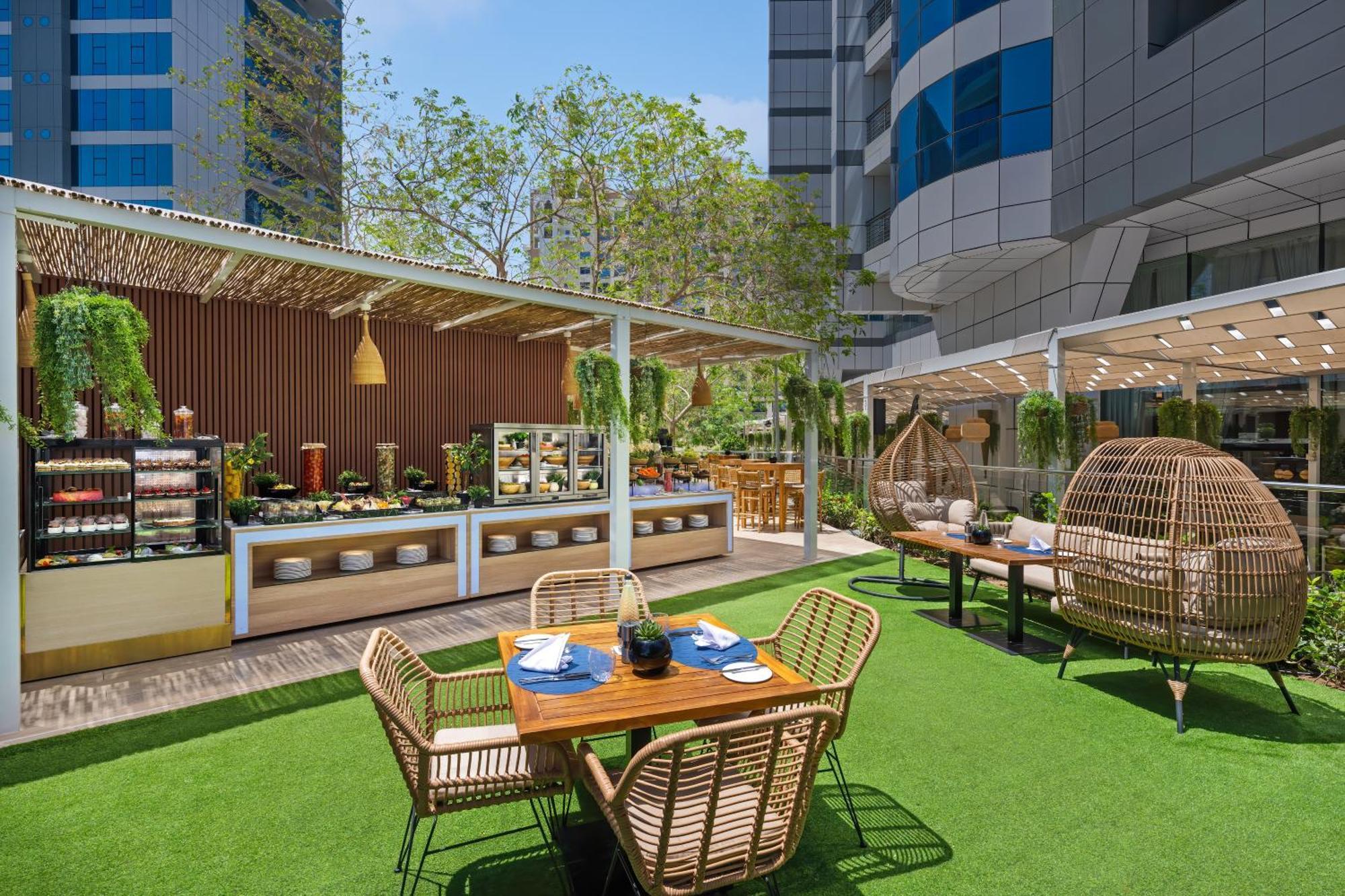 Dukes The Palm, A Royal Hideaway Hotel Dubai Exterior foto The photo shows a modern outdoor dining area, likely part of a restaurant or cafe. There are several seating arrangements including a wooden table surrounded by four wicker chairs. On the table, there are blue plates and a decorative item in the cent