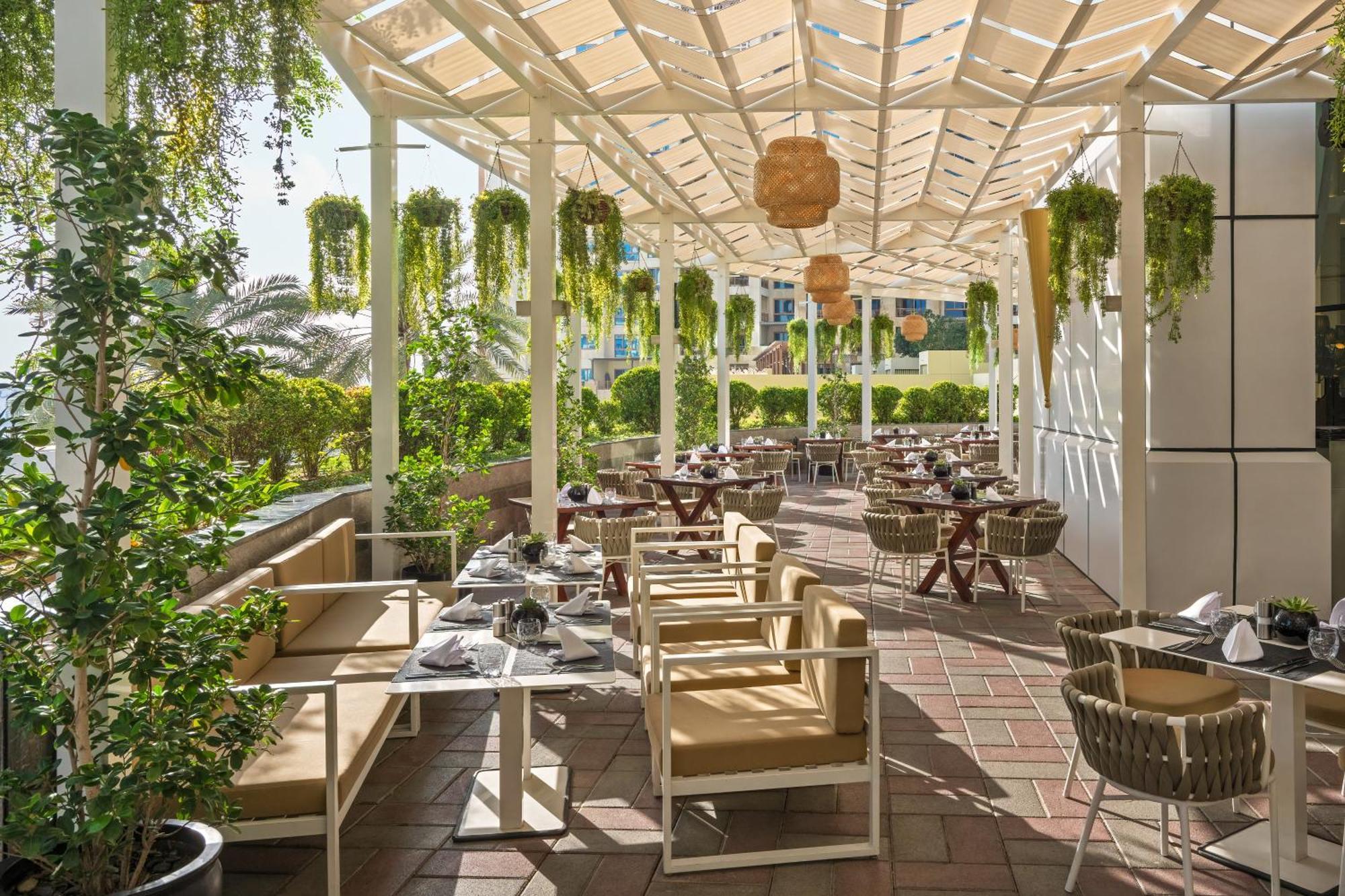 Dukes The Palm, A Royal Hideaway Hotel Dubai Exterior foto The photo shows an outdoor restaurant patio area with a bright and inviting atmosphere. There are several tables and chairs arranged neatly under a structured canopy. The furniture appears to be a mix of light-colored wooden pieces and wicker chairs.