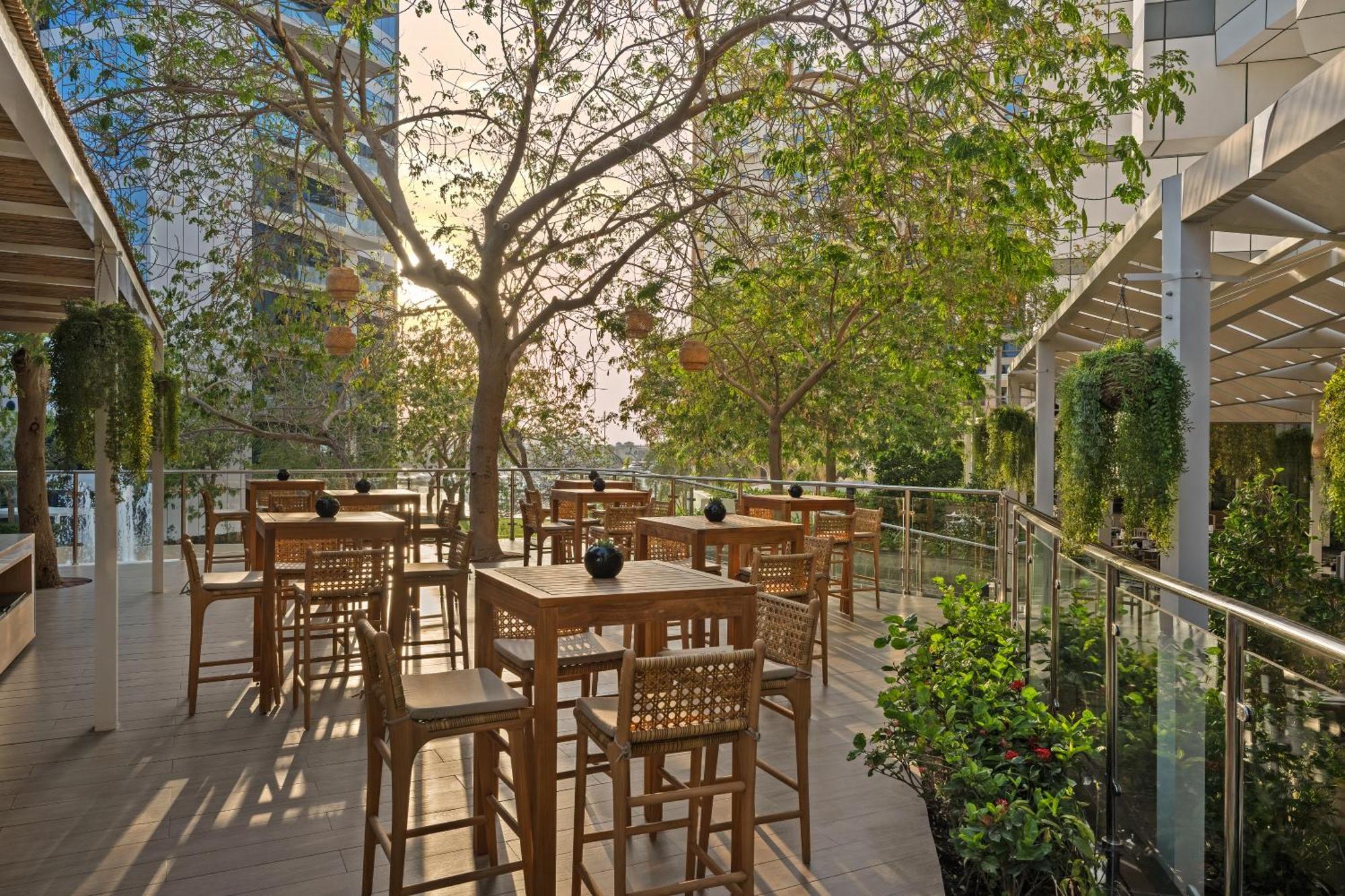 Dukes The Palm, A Royal Hideaway Hotel Dubai Exterior foto The photo shows a spacious outdoor dining area featuring wooden tables and chairs. There is a large tree with green leaves that provides shade, and the ambiance is enhanced by natural light filtering through the leaves. Surrounding the area are glass