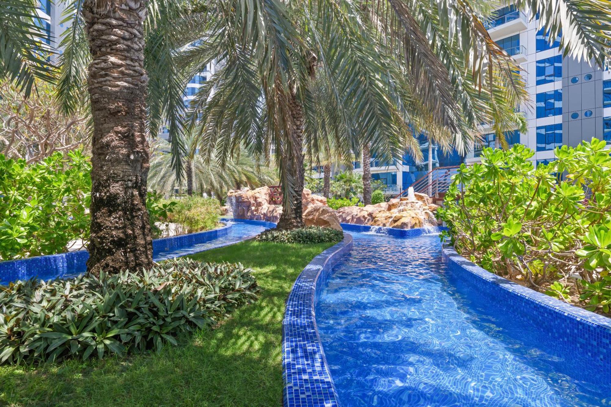 Dukes The Palm, A Royal Hideaway Hotel Dubai Exterior foto The photo depicts a beautifully landscaped tropical area featuring a winding waterway with clear blue water, bordered by lush greenery and palm trees. The vibrant blue tiles lining the water add a decorative touch, while the overall setting suggests 