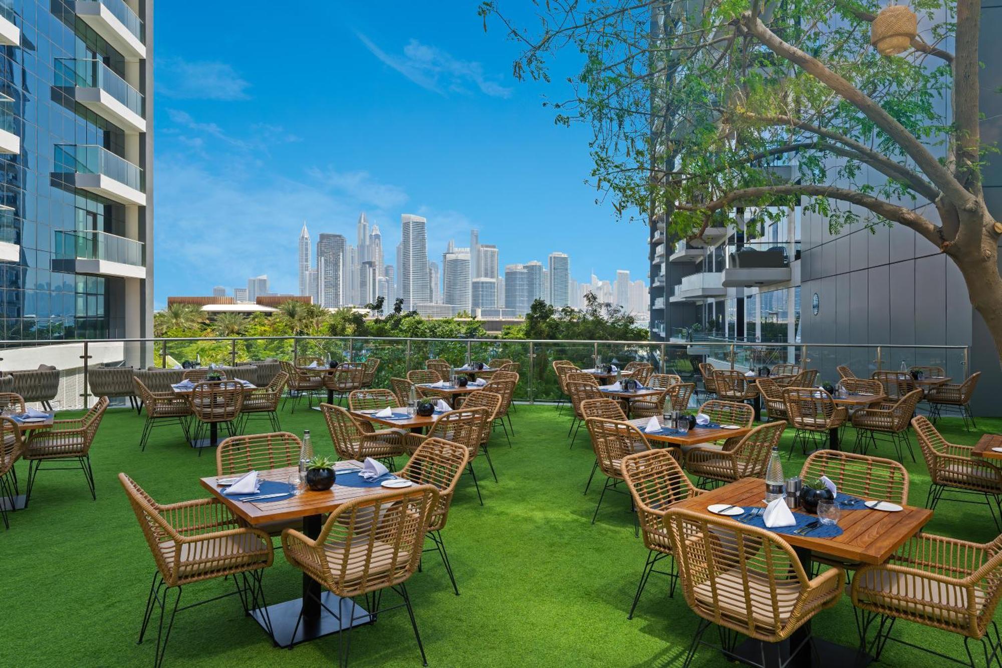 Dukes The Palm, A Royal Hideaway Hotel Dubai Exterior foto The photo shows an outdoor dining area with several wooden tables and wicker chairs arranged neatly. The tables are set with napkins and utensils, indicating readiness for guests. The dining area is surrounded by a green lawn and features a tree, pro