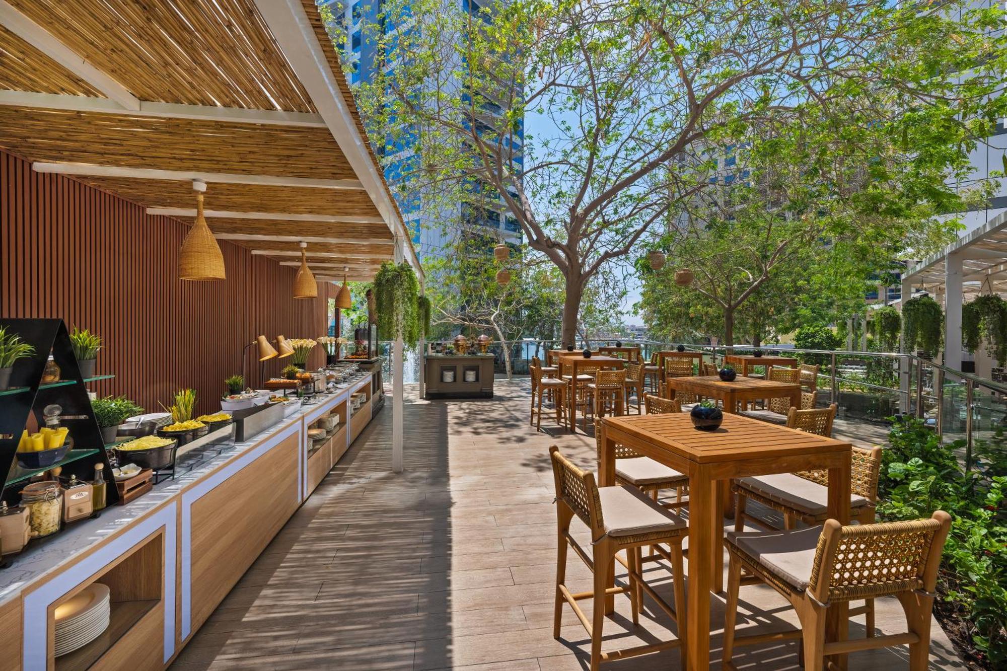 Dukes The Palm, A Royal Hideaway Hotel Dubai Exterior foto The photo showcases an outdoor dining area in a restaurant or café. It features a wooden terrace with several tables and chairs arranged for patrons. On one side, there is a buffet or serving area with various displays of food items. 

Surrounding th
