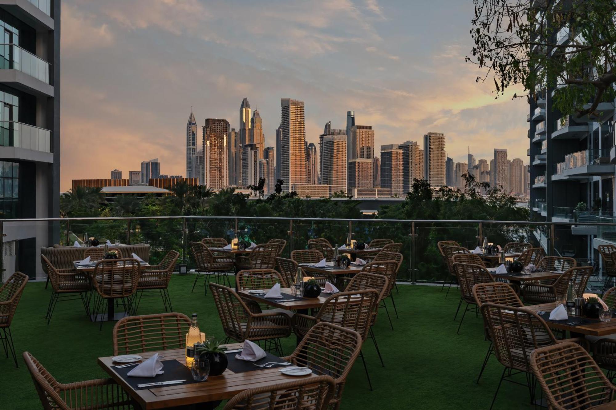 Dukes The Palm, A Royal Hideaway Hotel Dubai Exterior foto The photo shows an outdoor dining area with several tables and chairs arranged neatly. The tables are set with plates and candles, creating an inviting atmosphere. In the background, there is a stunning city skyline featuring tall skyscrapers, bathed