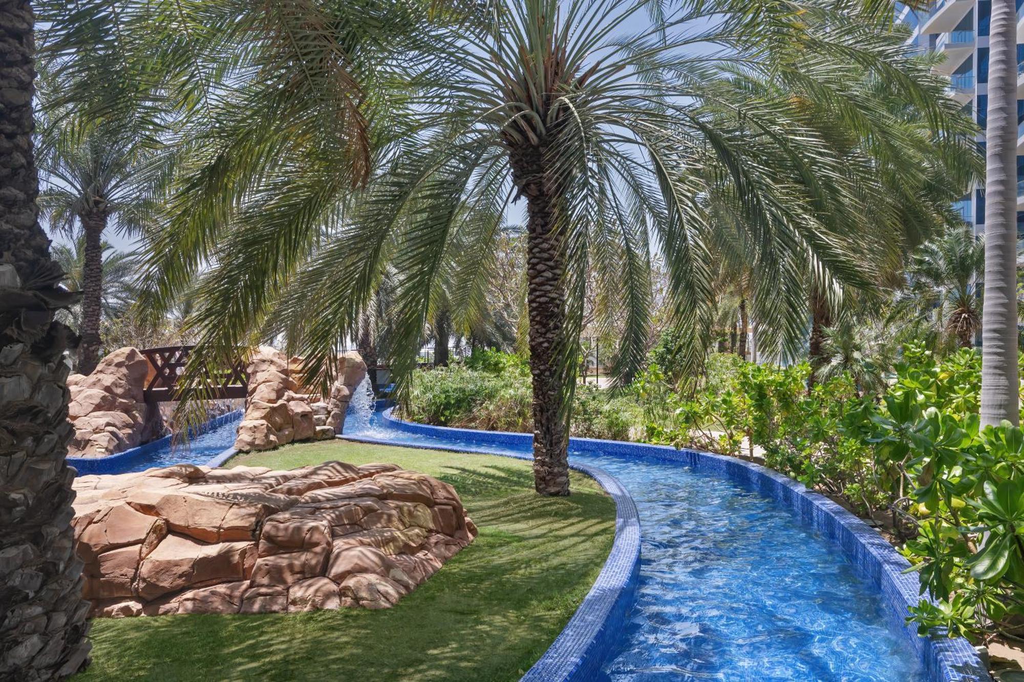 Dukes The Palm, A Royal Hideaway Hotel Dubai Exterior foto The photo depicts a serene outdoor setting, featuring a winding blue water channel surrounded by lush greenery. Palm trees tower over the scene, providing shade and a tropical feel. There are rocky formations integrated into the landscape, and the ov