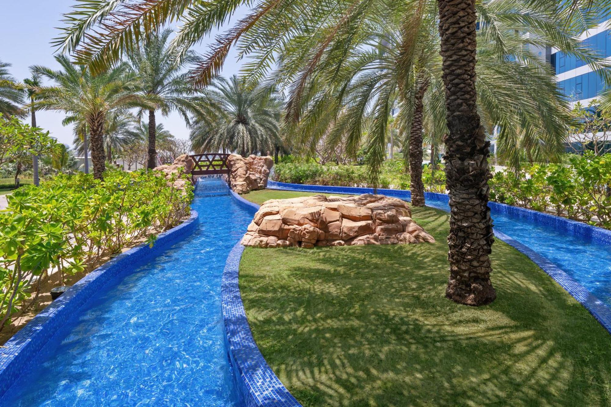 Dukes The Palm, A Royal Hideaway Hotel Dubai Exterior foto The photo depicts a serene outdoor area featuring a winding, clear blue waterway surrounded by lush greenery and palm trees. In the background, there is a small stone bridge arching over the water. Natural rock formations and manicured grass create a