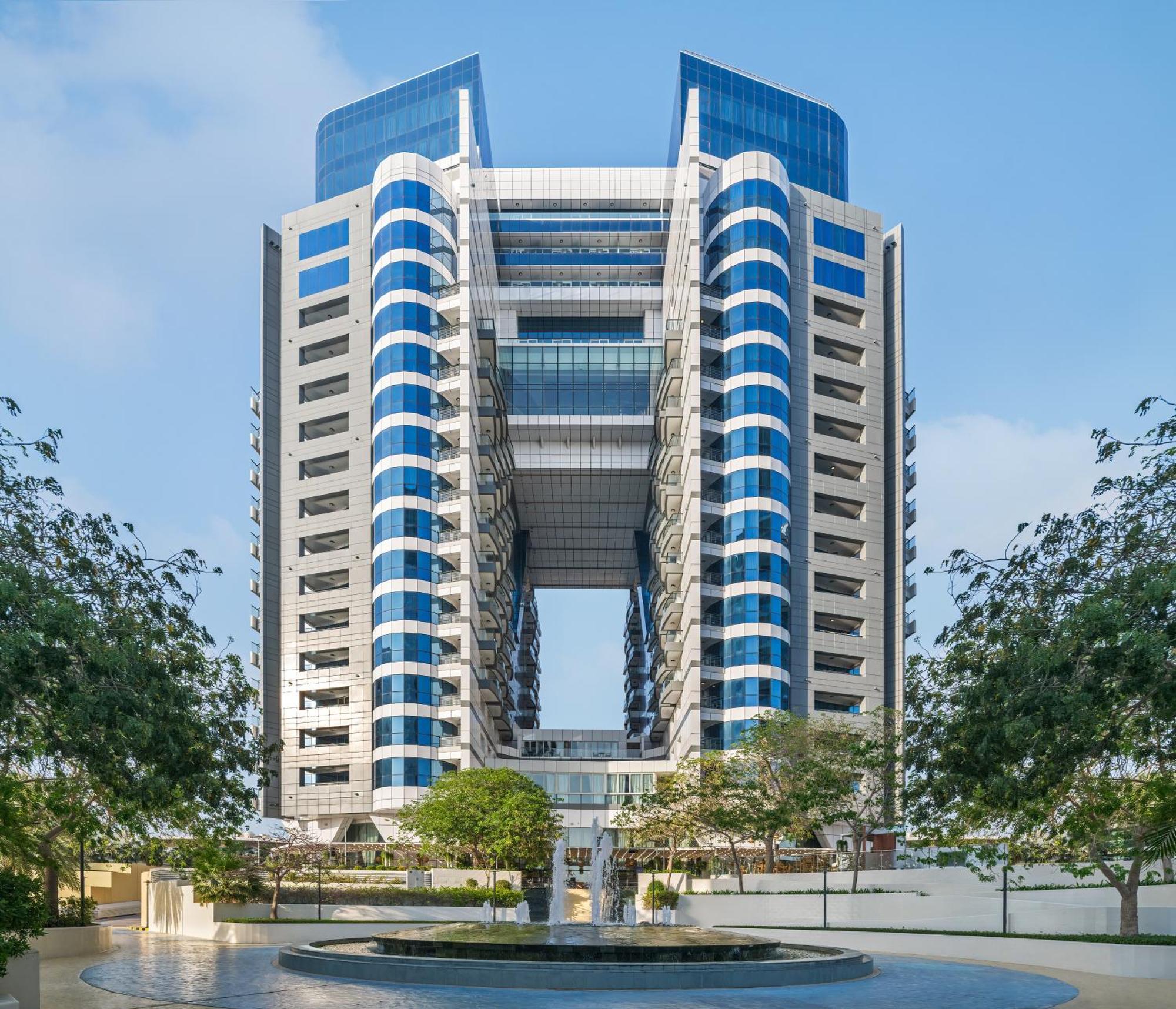 Dukes The Palm, A Royal Hideaway Hotel Dubai Exterior foto The image shows a large modern building characterized by a distinctive architectural design. It features a symmetrical façade with multiple levels and large, blue-tinted glass windows. The structure has prominent vertical elements and an open center,