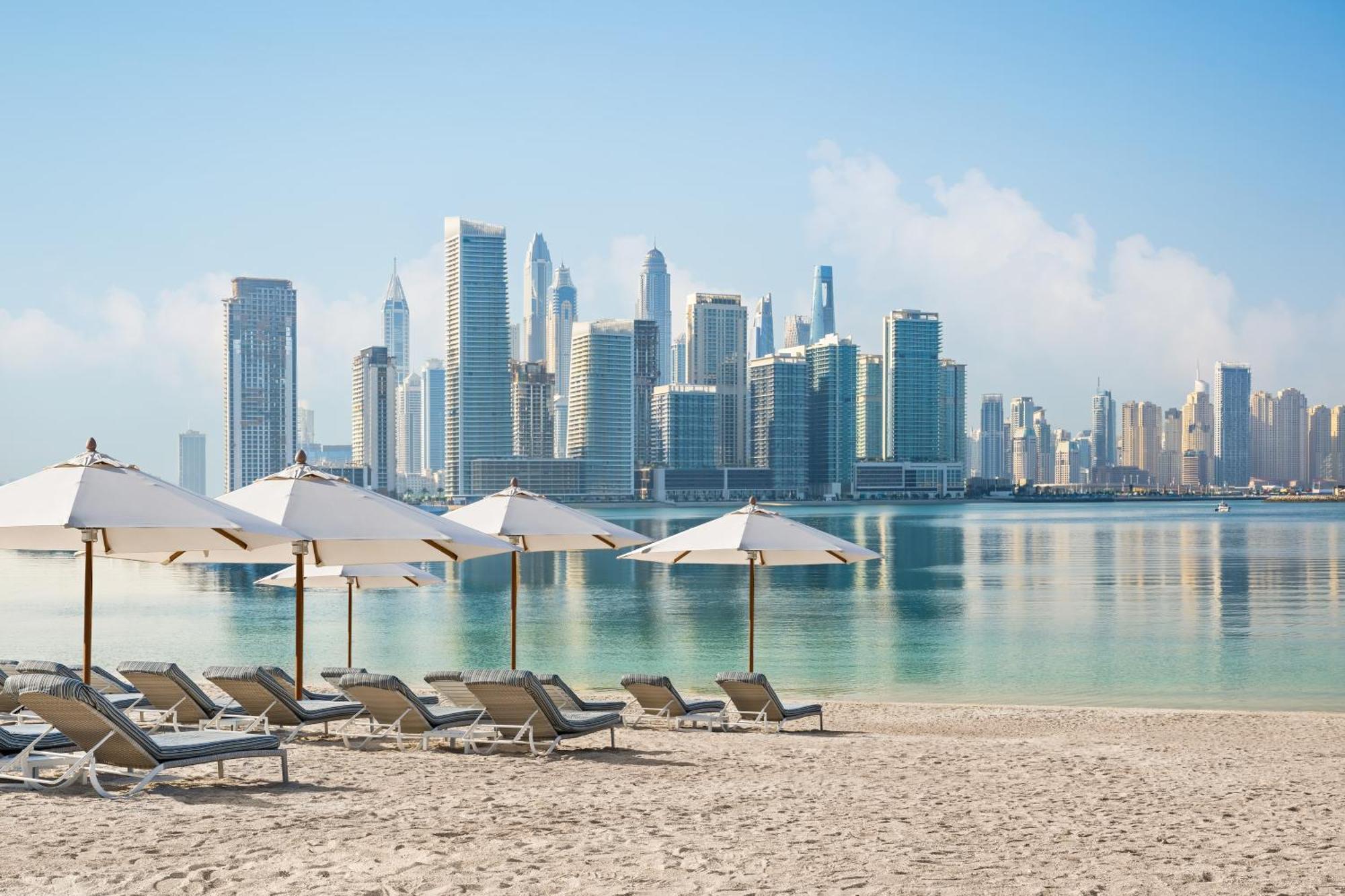 Dukes The Palm, A Royal Hideaway Hotel Dubai Exterior foto The photo showcases a serene beach scene with several lounge chairs positioned on the sandy shore. Each chair is paired with a large umbrella for shade. In the background, a modern city skyline is visible, featuring tall skyscrapers and a clear blue 