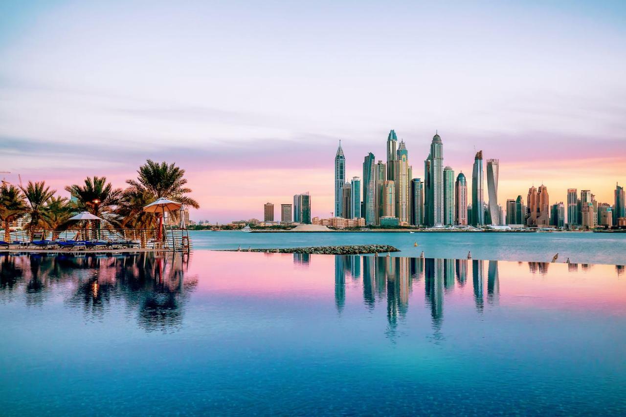 Dukes The Palm, A Royal Hideaway Hotel Dubai Exterior foto The image showcases a stunning skyline view of a modern city, likely during sunrise or sunset, as indicated by the beautiful pastel colors in the sky. The foreground features a calm body of water, reflecting the skyscrapers and the colorful sky. Lush